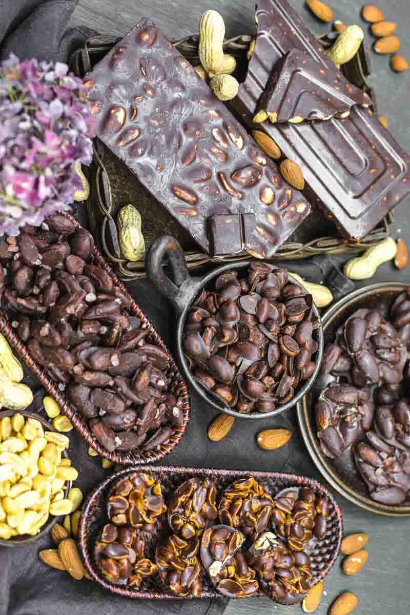 An assortment of dark chocolate bars and clusters with peanuts and almonds, arranged in various baskets and bowls on a dark surface.