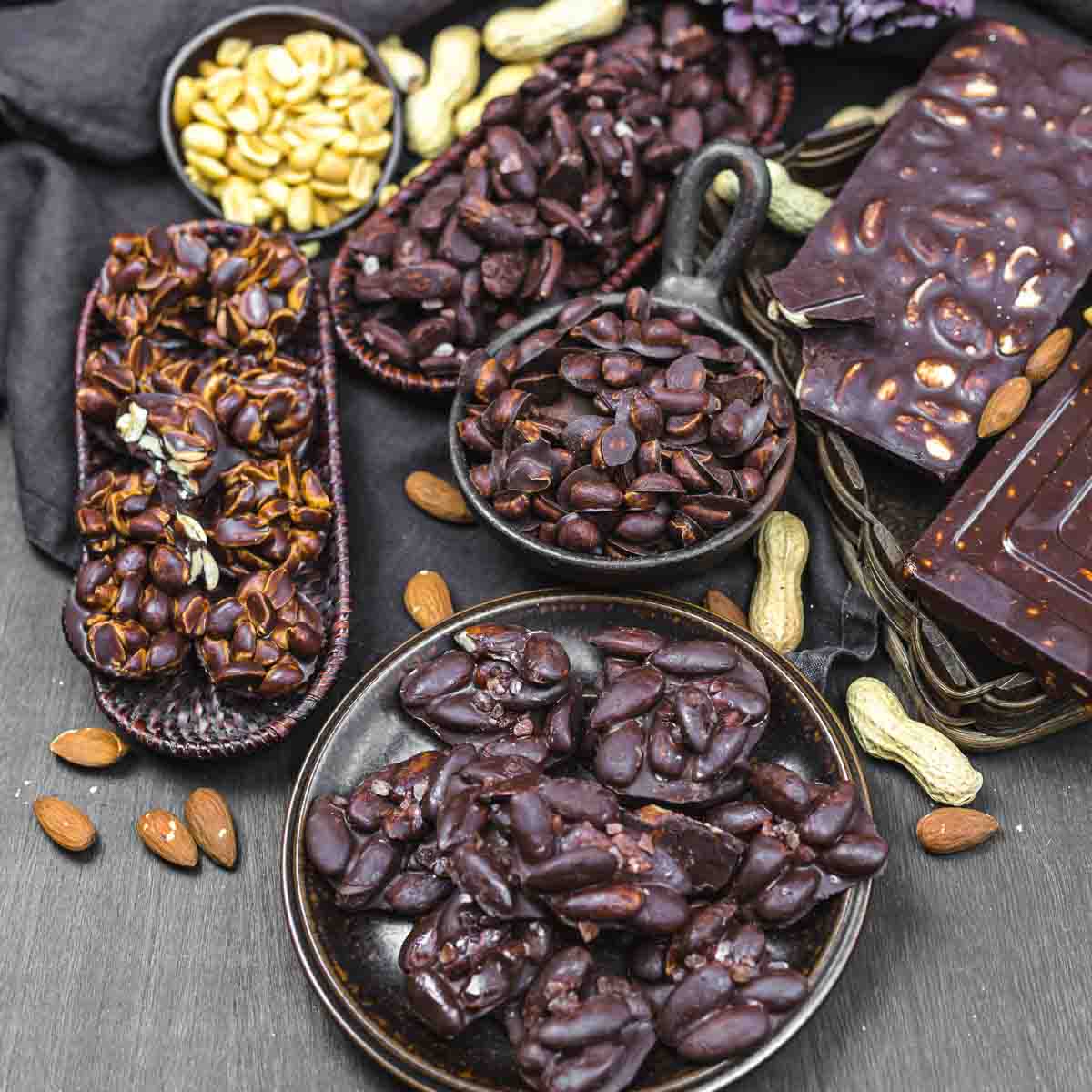 Various chocolate-covered nuts, including peanuts and almonds, arranged in bowls and trays on a dark surface. Whole almonds and peanuts scattered around.