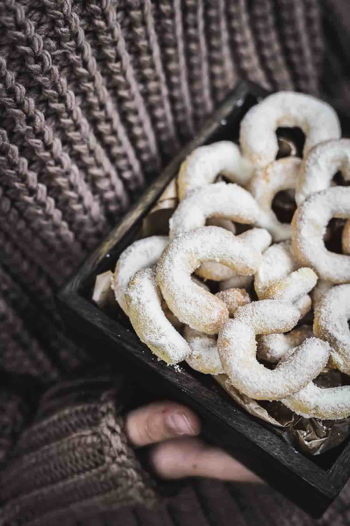 A person in a knitted sweater holds a box of powdered sugar-covered crescent cookies.