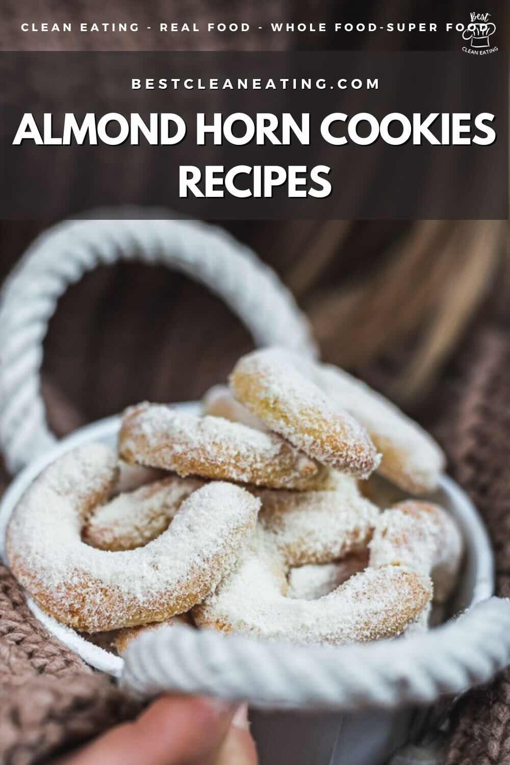 A person holds a white bowl filled with almond horn cookies dusted with powdered sugar. Text at the top reads: "BESTCLEANEATING.COM ALMOND HORN COOKIES RECIPES.