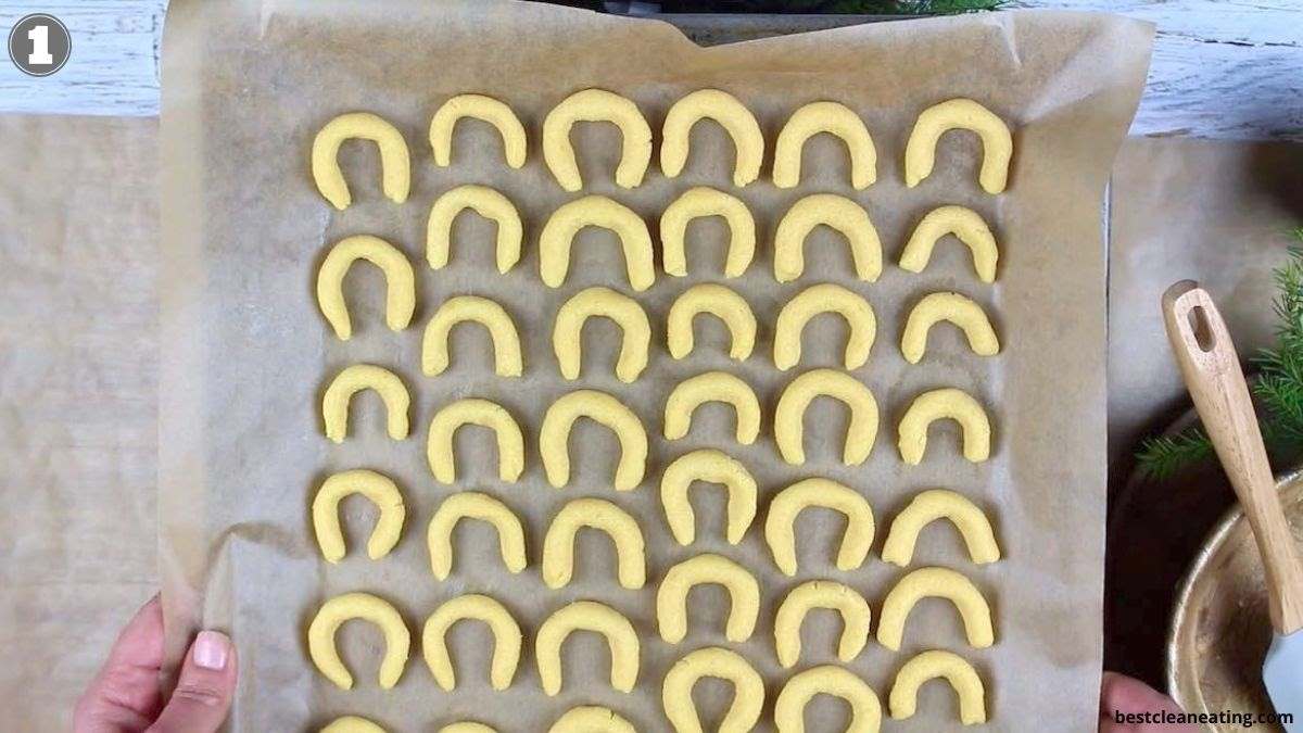 A baking tray with rows of unbaked crescent-shaped cookies on parchment paper.