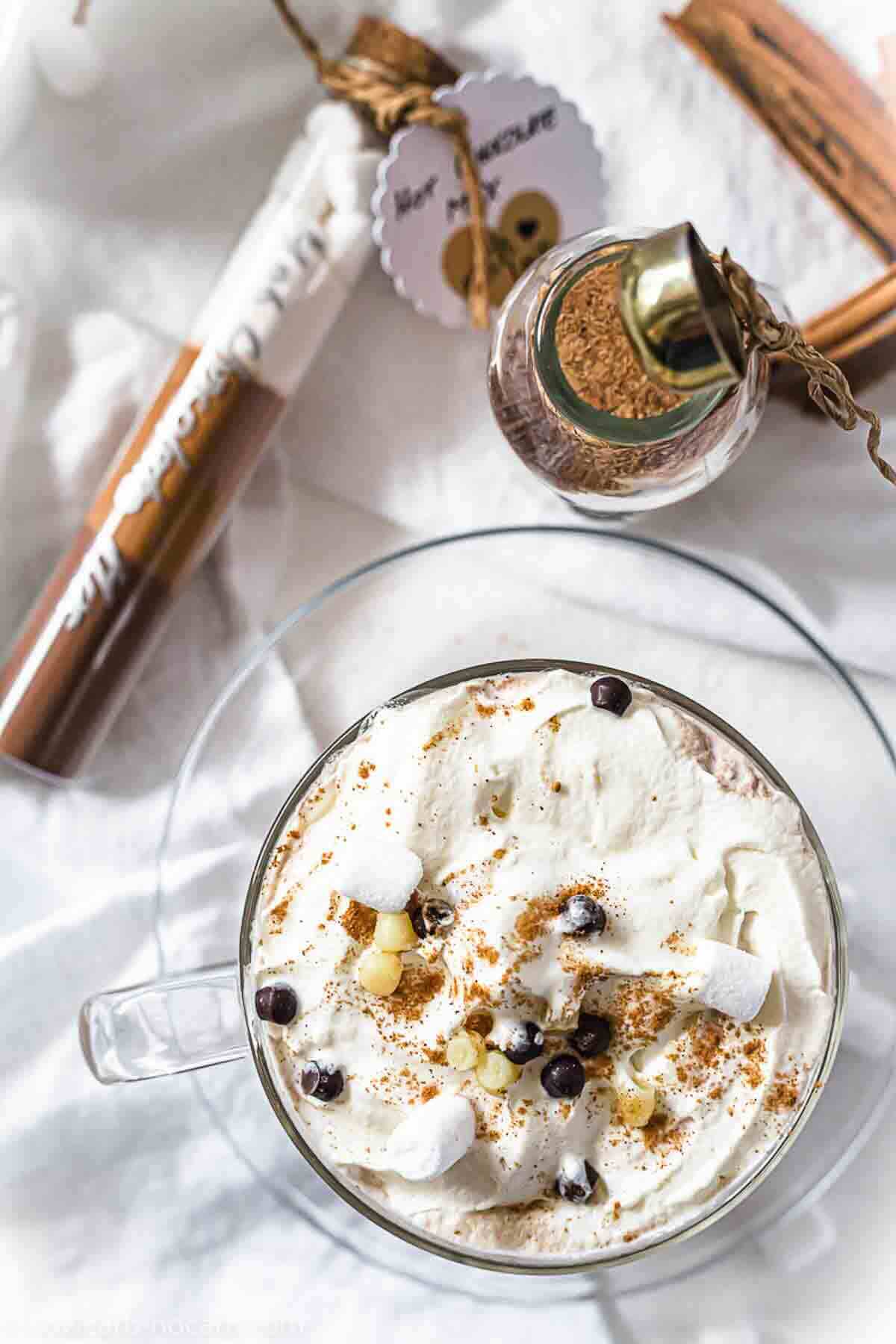 A clear mug filled with a creamy beverage topped with whipped cream, chocolate chips, and mini marshmallows. Nearby are a jar of spices and a chocolate wafer stick.