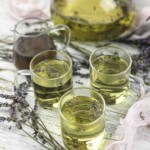 Three glass mugs of lavender tea on a white wooden surface, surrounded by lavender sprigs. A glass jug with more tea is in the background.