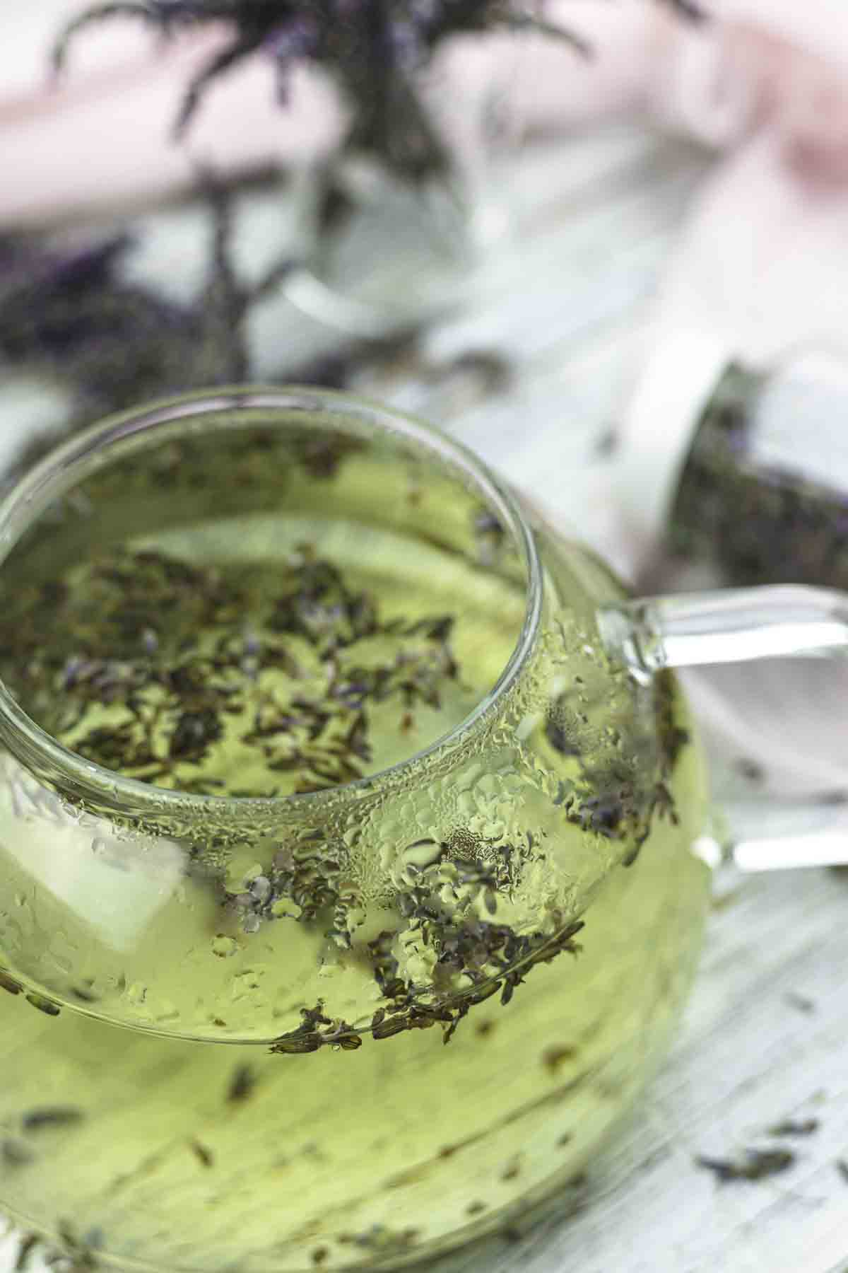 A glass teapot filled with light green liquid, containing floating lavender petals, sits on a wooden surface with scattered lavender around it.