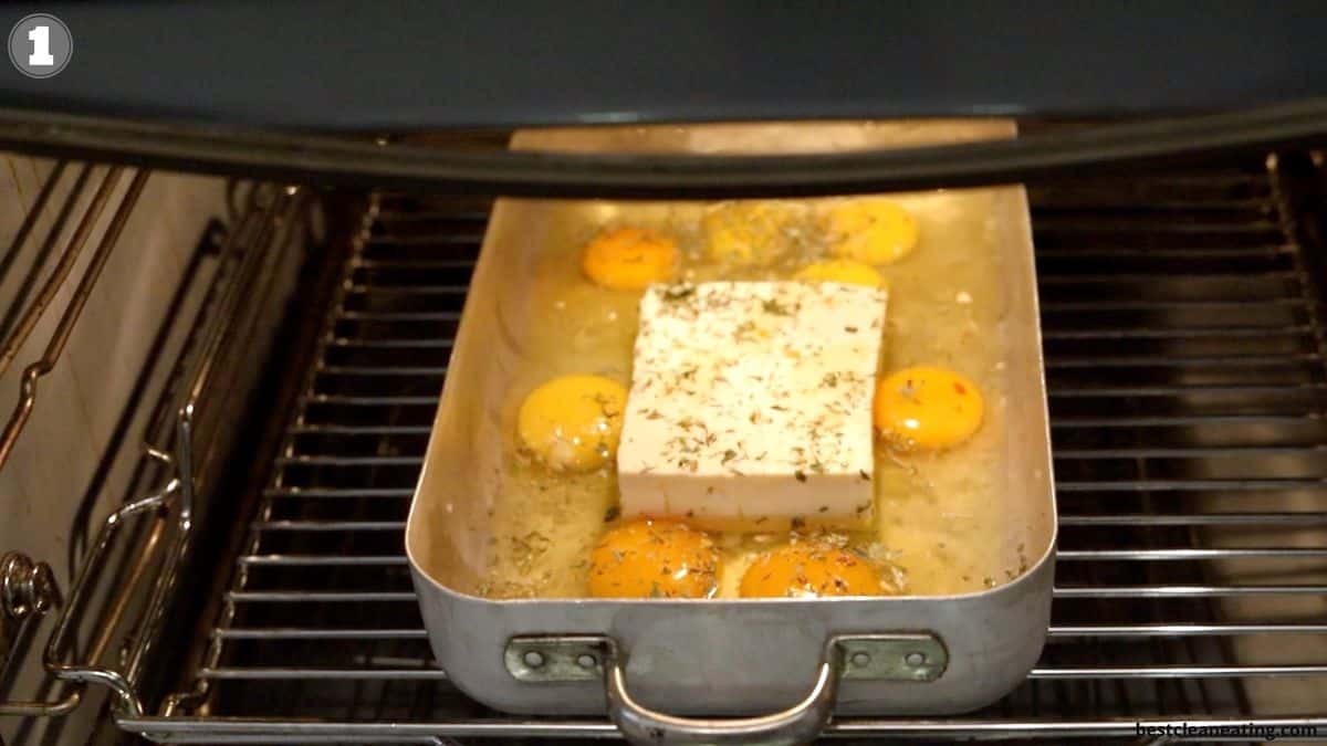 A metal pan in an oven contains eggs, a block of cheese, and herbs, all uncooked.