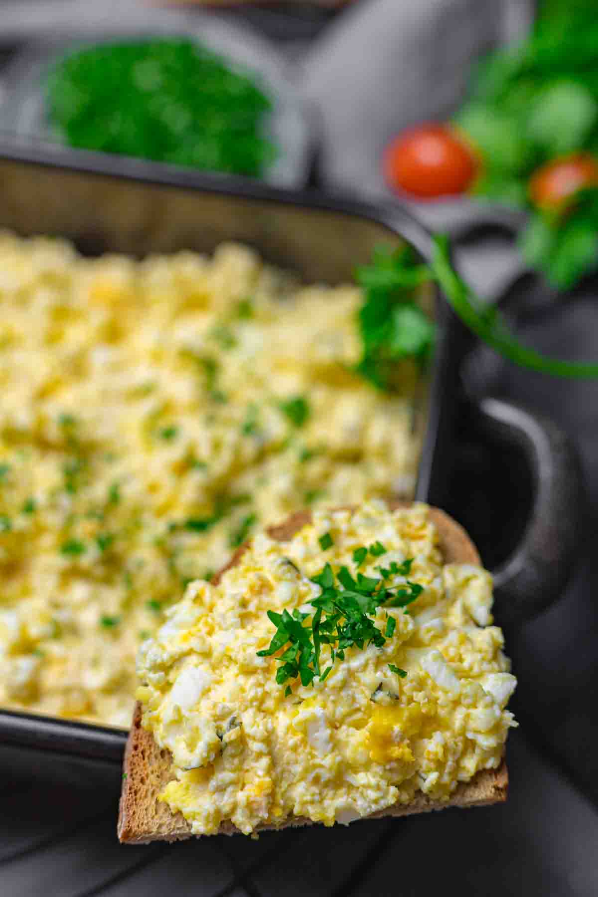 Egg salad on a slice of bread garnished with parsley, with a baking dish of more egg salad and blurred vegetables in the background.