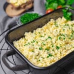 A black rectangular dish filled with scrambled eggs, garnished with chopped parsley. In the background, there are slices of bread and a bowl of fresh greens.
