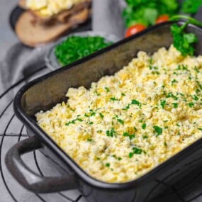 A black rectangular dish filled with scrambled eggs, garnished with chopped parsley. In the background, there are slices of bread and a bowl of fresh greens.