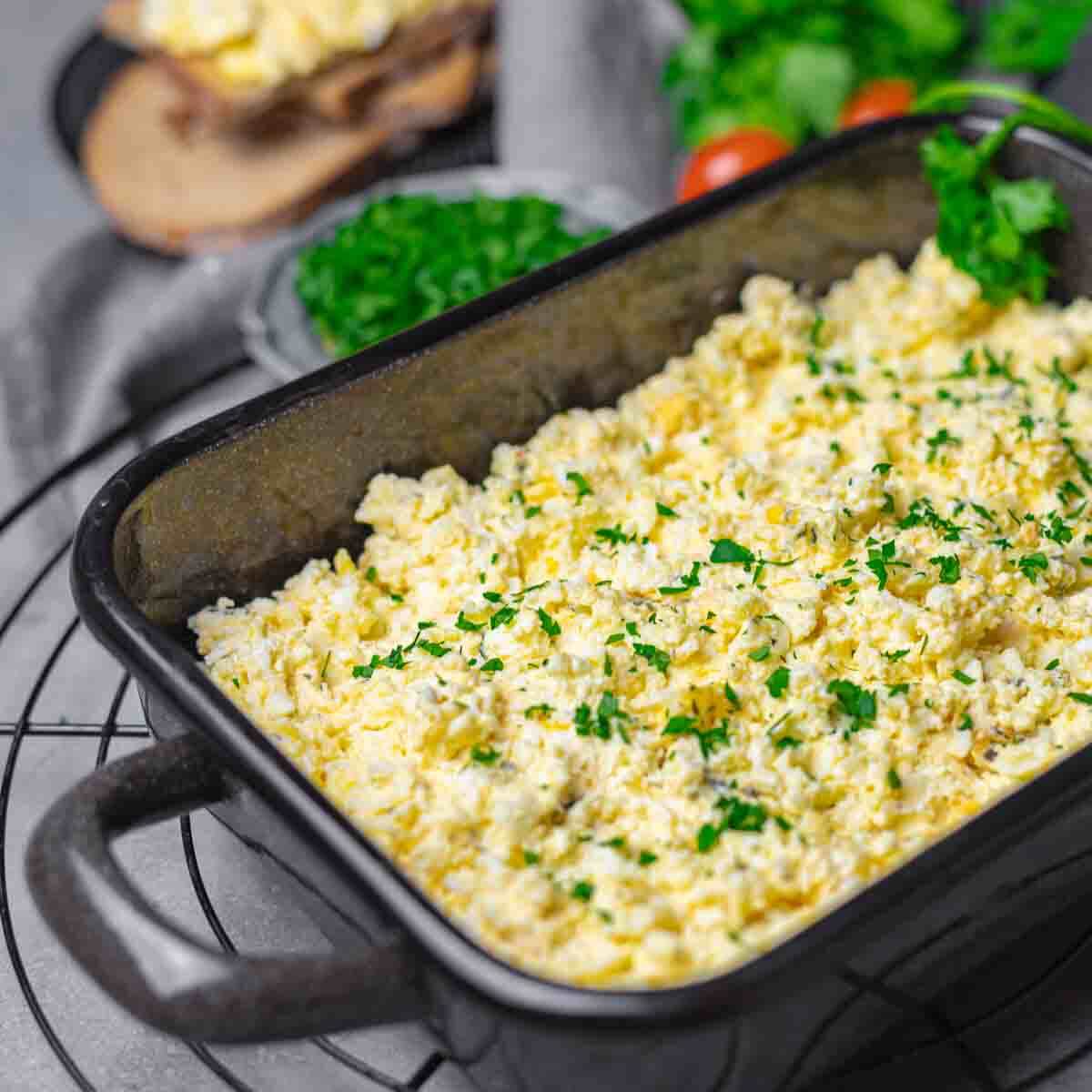 A black rectangular dish filled with scrambled eggs, garnished with chopped parsley. In the background, there are slices of bread and a bowl of fresh greens.