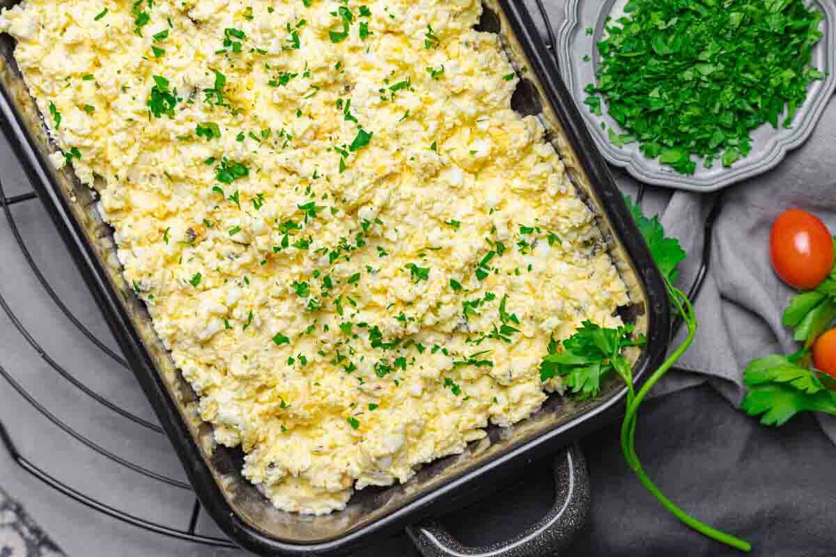 Baking dish with eggs and feta casserole topped with chopped parsley, next to a bowl of parsley and cherry tomatoes.