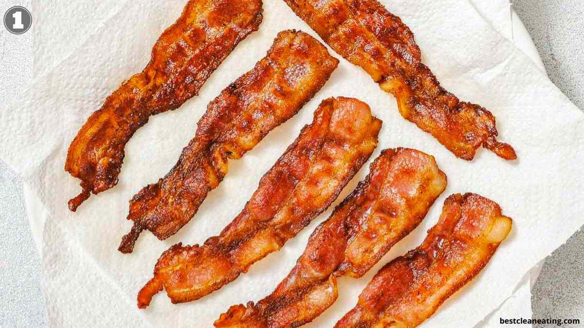 Six cooked strips of bacon are laid out on a white paper towel, likely to absorb excess grease.