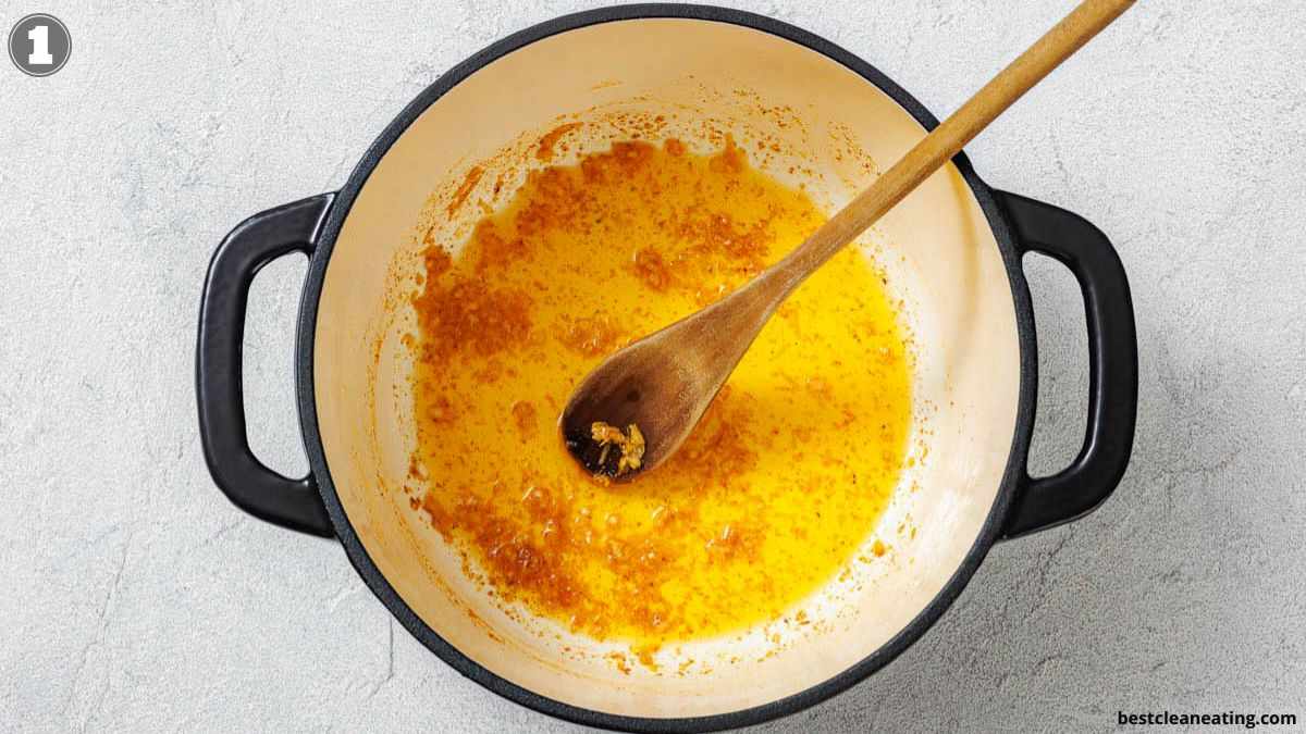 A wooden spoon rests in a white pot with black handles, containing sautéed onions and spices in oil on a light surface.