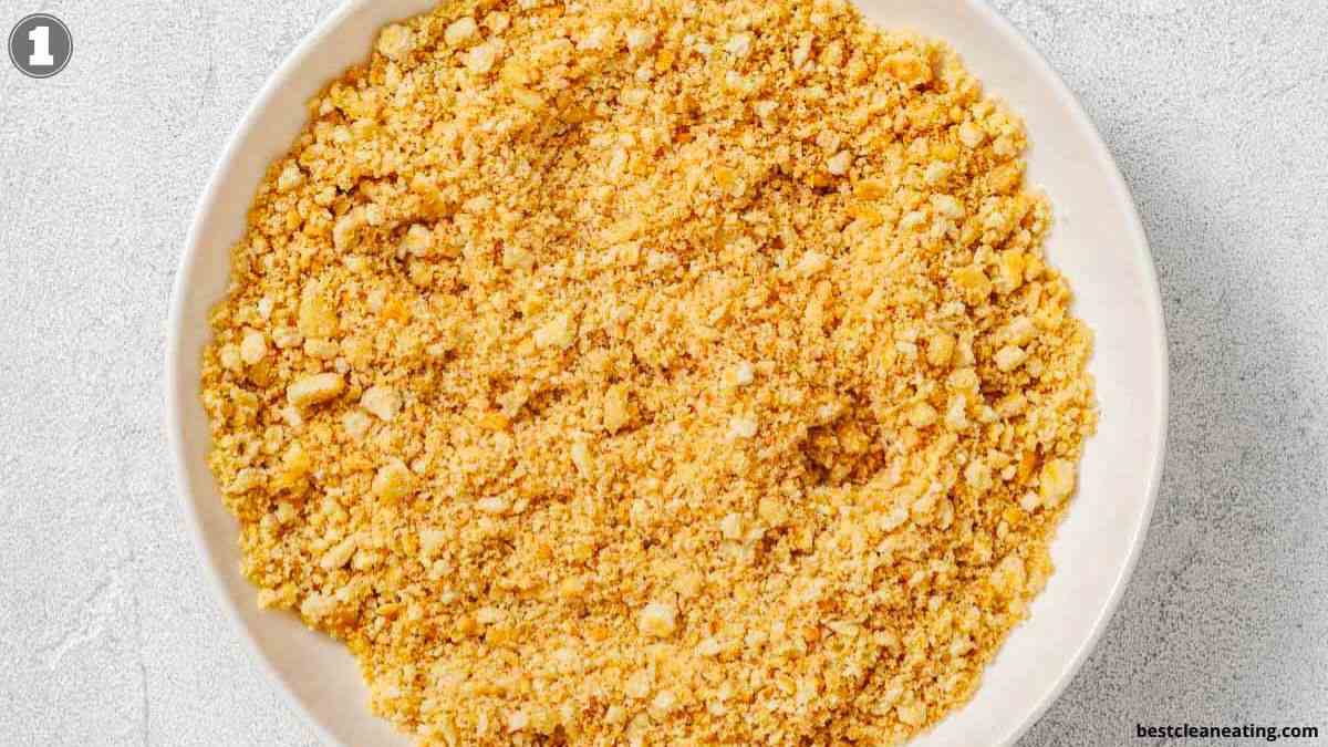 A white bowl filled with finely crushed cornflakes or breadcrumbs seen from above on a light surface.