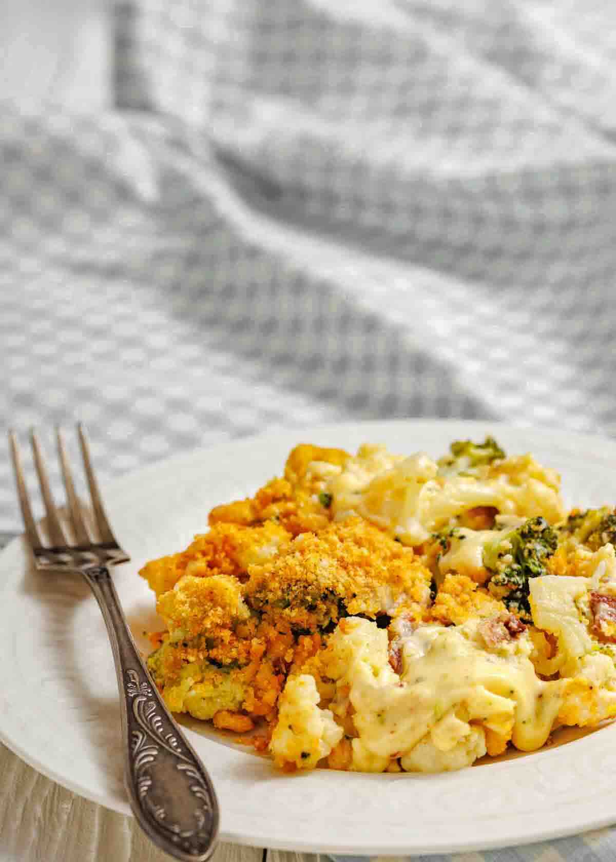 A plate with a serving of broccoli and cheese casserole topped with breadcrumbs, next to a fork on a white surface.
