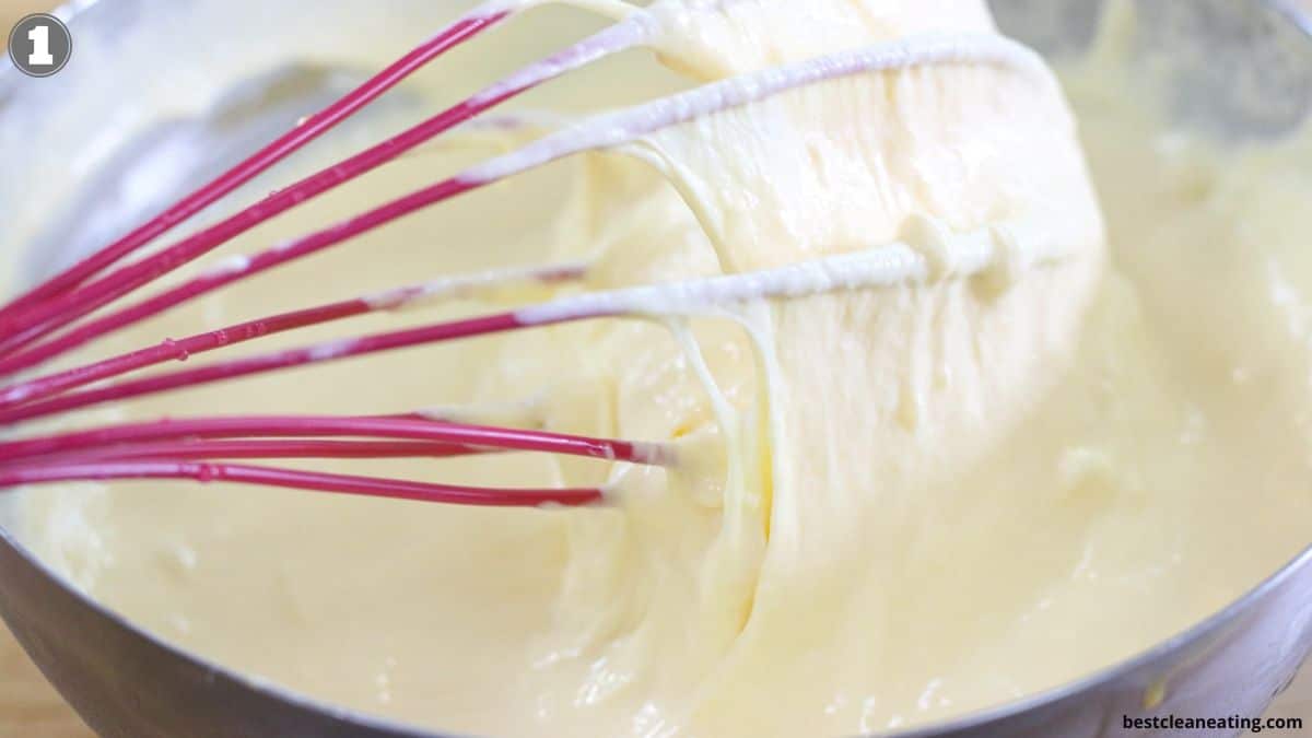 A red whisk mixes thick, creamy batter in a stainless steel bowl.
