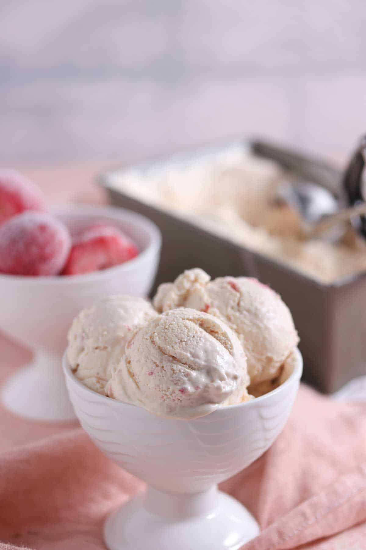 A white bowl filled with scoops of ice cream sits in the foreground, with a bowl of frozen strawberries and a container of ice cream with a scoop in the background.