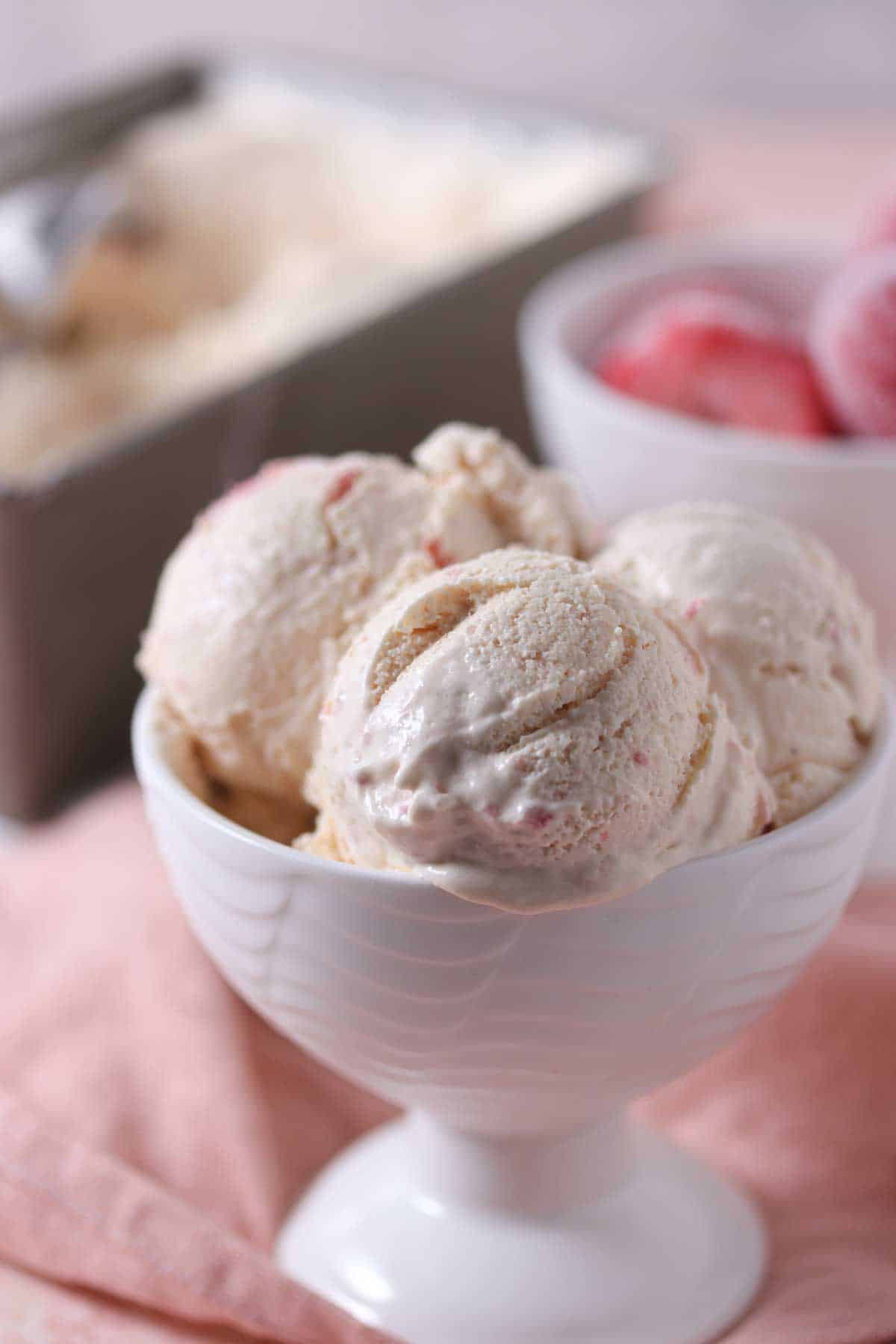 Three scoops of strawberry ice cream in a white bowl are placed on a pink cloth, with a container and another bowl of ice cream in the background.