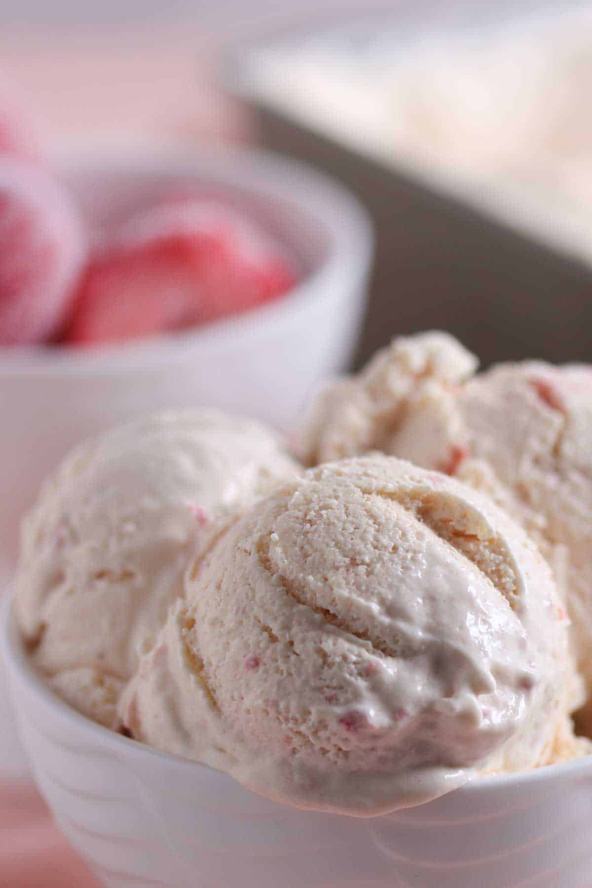 A bowl of strawberry ice cream with visible pink bits, with a blurred container of strawberries in the background.