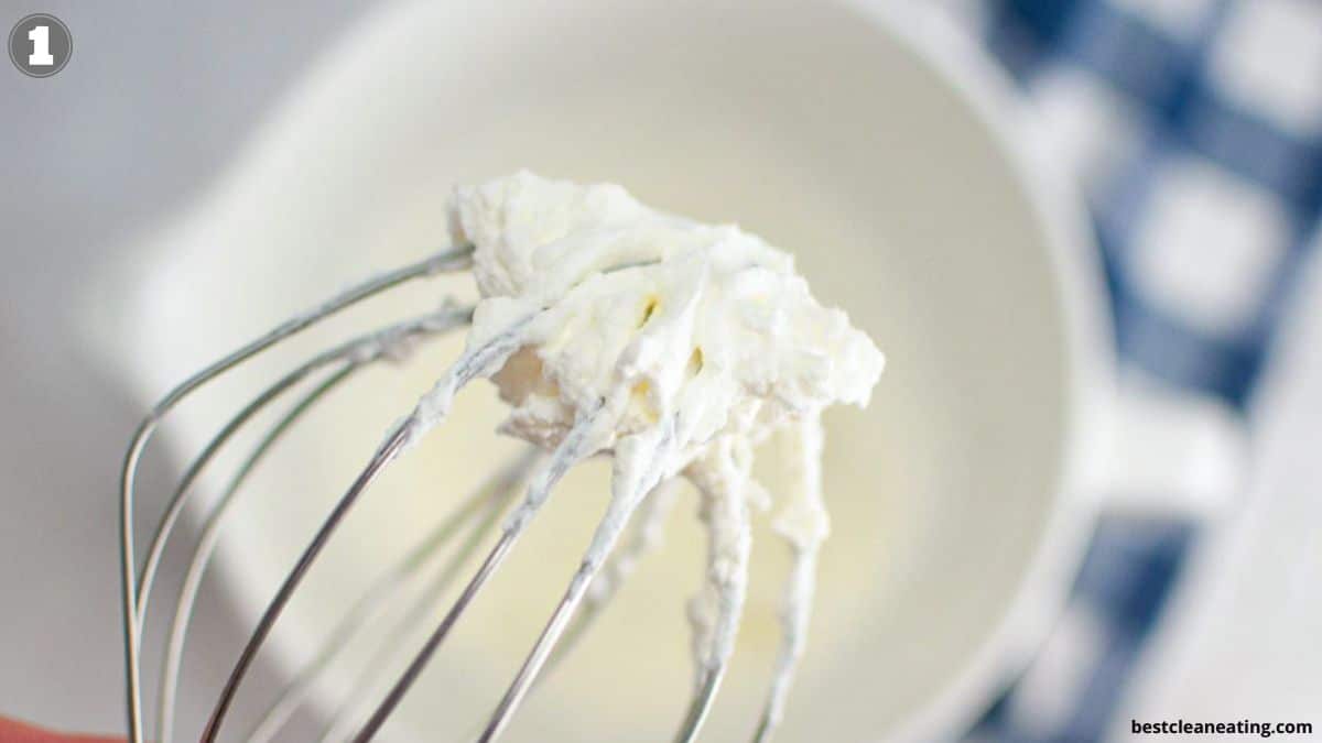 A close-up of a metal whisk with freshly whipped cream, with a white bowl and blue checkered cloth in the blurred background.