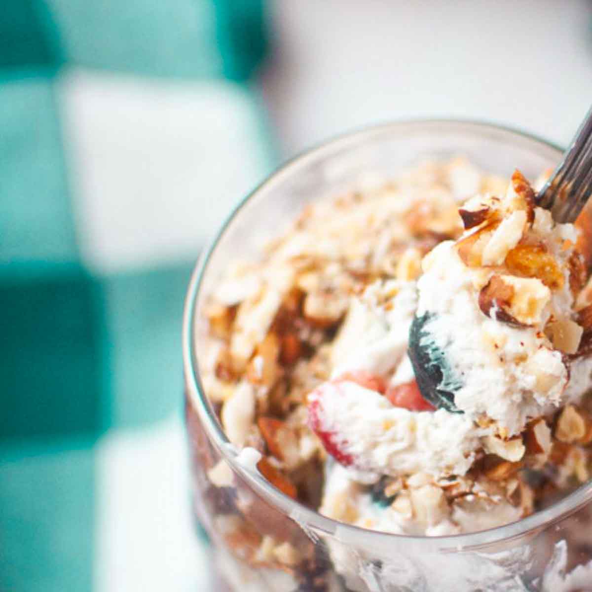 A close-up of a spoonful of parfait with whipped cream, berries, and granola in a glass, with a teal and white checkered cloth in the background.
