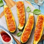 Four baked zucchini boats filled with melted cheese and tomato mixture, served on parchment paper with basil leaves and a side of tomato sauce.