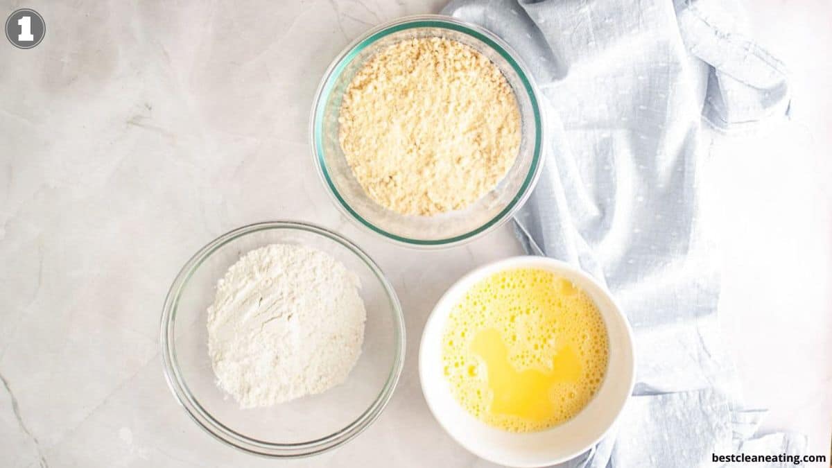 Three bowls containing whisked eggs, flour, and breadcrumbs are arranged on a light surface next to a folded light blue cloth.