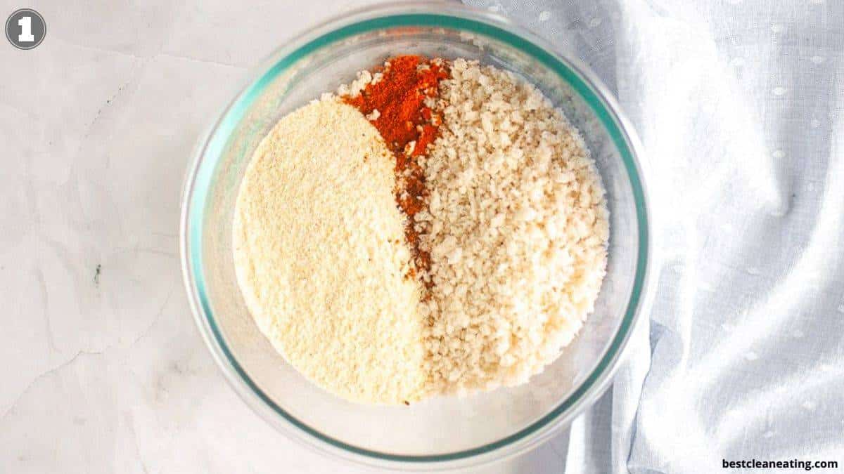 A glass bowl containing three separate piles of dry ingredients: breadcrumbs, almond flour, and paprika, on a light surface with a blue cloth nearby.