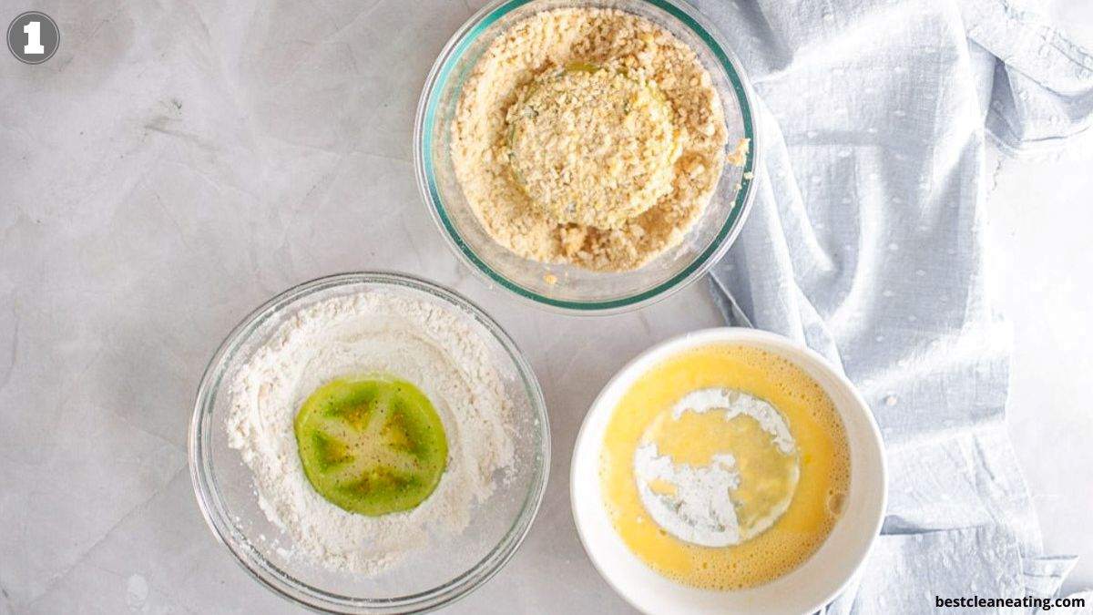 Three bowls on a white surface: one with flour, one with beaten eggs, and one with breadcrumbs, each containing a slice of green tomato being coated.