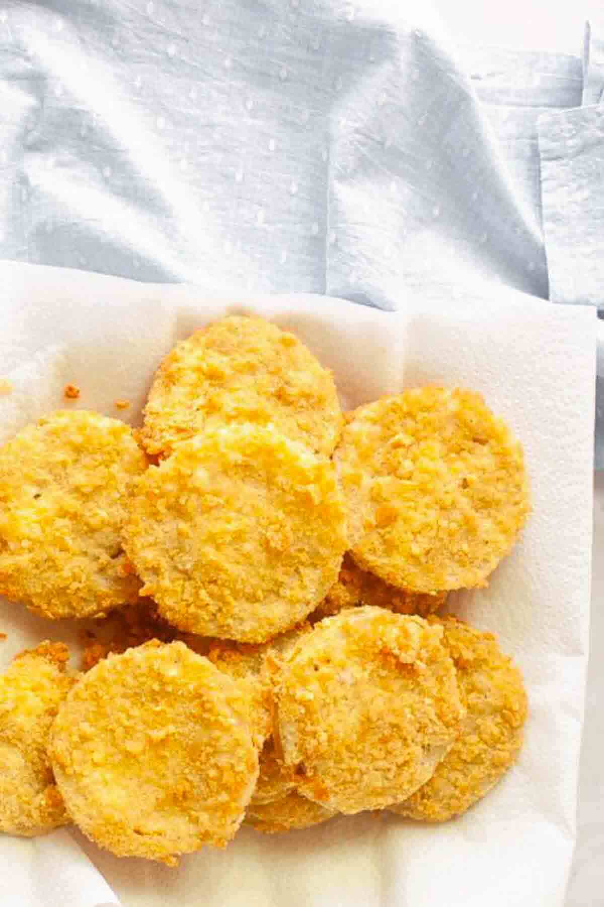 Golden, breaded round slices of fried green tomatoes arranged on a white paper towel, with a light blue textured cloth in the background.