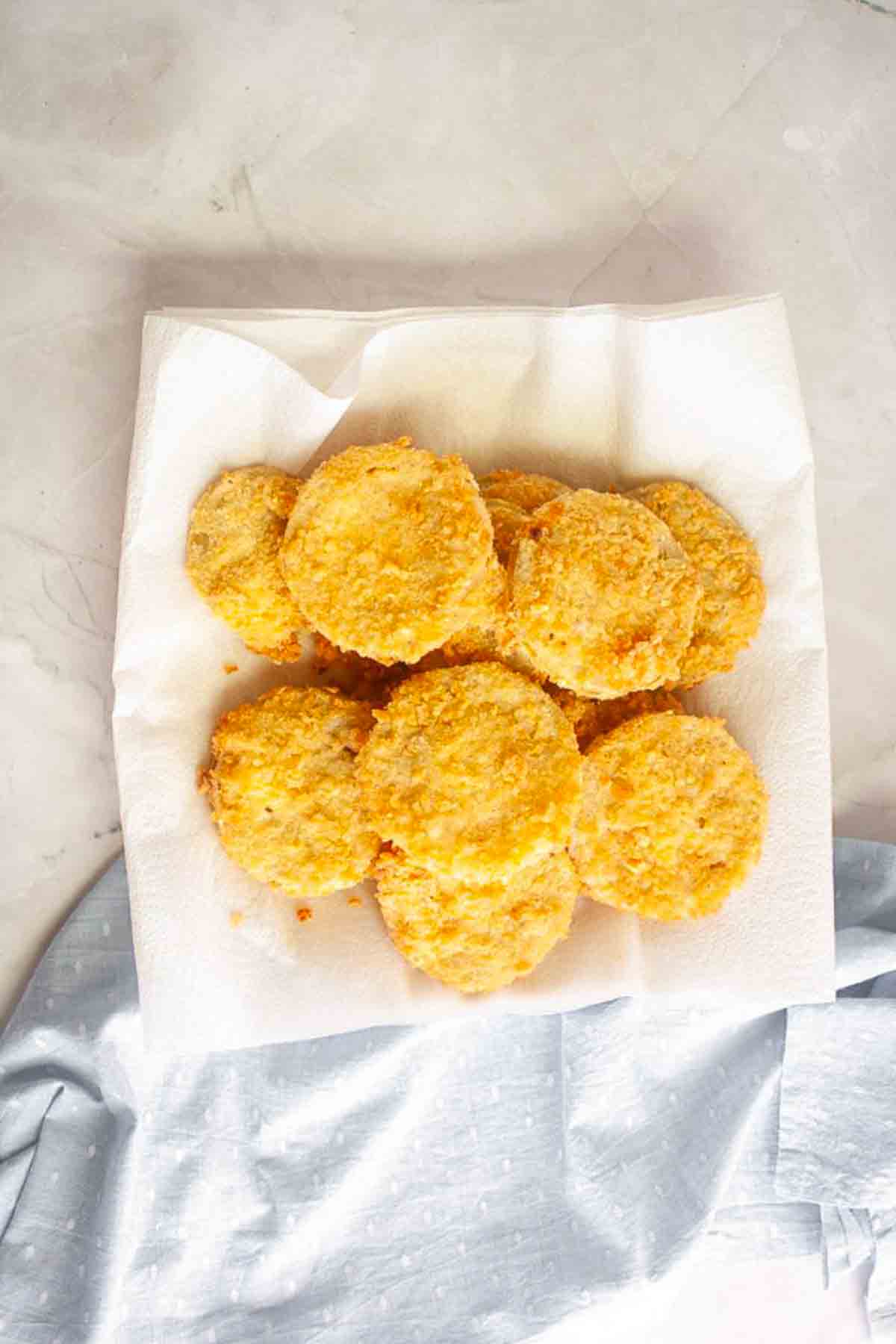A stack of golden, round fried green tomatoes sits on a white paper towel atop a light-colored surface with part of a blue cloth visible.