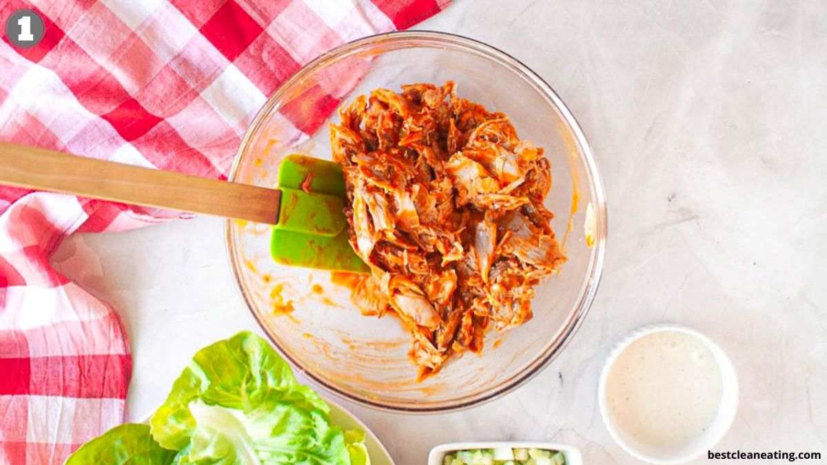A glass bowl with shredded chicken mixed with sauce and a spatula, next to lettuce, chopped cucumbers, a cup of sauce, and a red checkered cloth on a white surface.