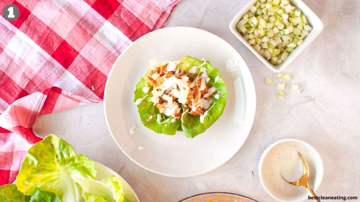 A lettuce wrap filled with diced chicken and white sauce on a plate, surrounded by a bowl of diced cucumbers, dressing, and a red checkered napkin.
