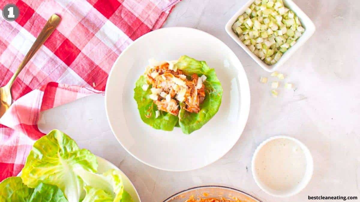 A lettuce leaf topped with shredded chicken and diced vegetables on a white plate, surrounded by lettuce, dressing, and a bowl of diced cucumbers on a light surface.