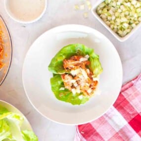 A lettuce leaf filled with shredded chicken and diced onions on a white plate, surrounded by diced cucumbers, lettuce, sauce, and a red plaid napkin.