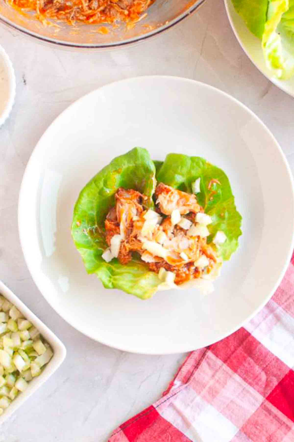 A white plate with a lettuce leaf wrap filled with shredded meat and chopped onions, on a light surface with a red checkered cloth nearby.