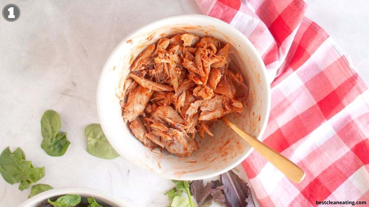 A bowl of shredded cooked chicken with a fork sits on a white surface next to a red plaid towel and some leafy greens.