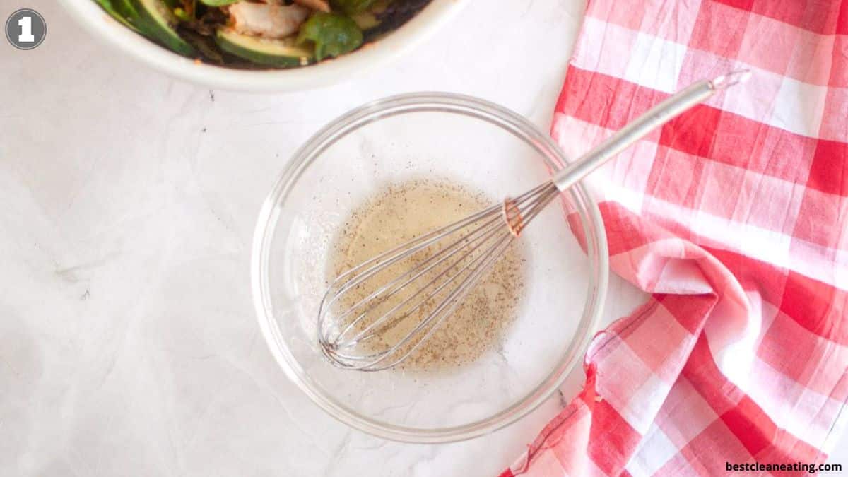 A clear glass bowl with a whisk and a small amount of liquid sits on a white surface next to a red and white checkered cloth.