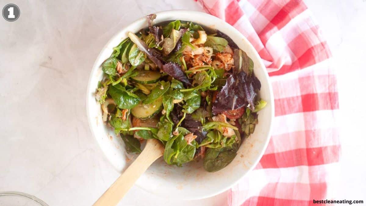 A white bowl filled with mixed green salad and a wooden spoon rests on a marble surface next to a red and white checkered cloth.