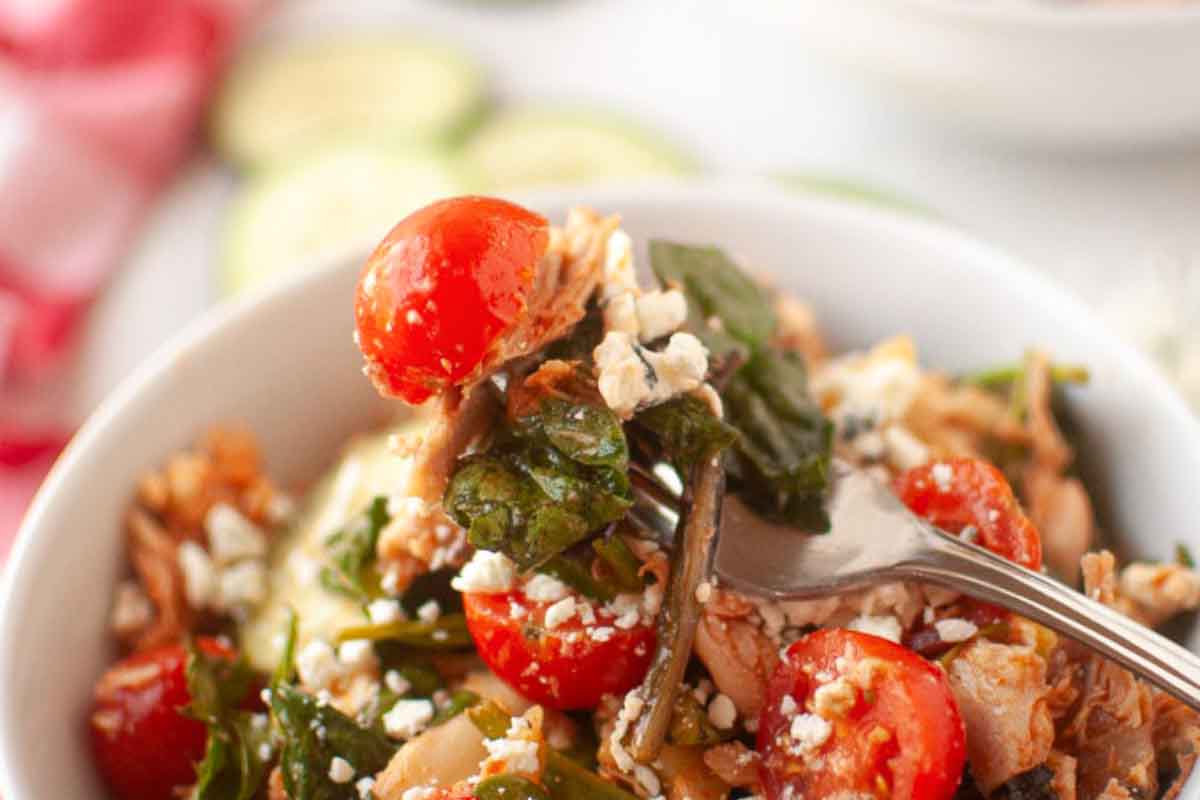 A close-up of a fork in a bowl of salad with cherry tomatoes, leafy greens, shredded chicken, and crumbled cheese.