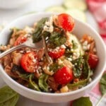 A bowl of salad with shredded meat, cherry tomatoes, spinach, and crumbled cheese, with a fork resting inside.