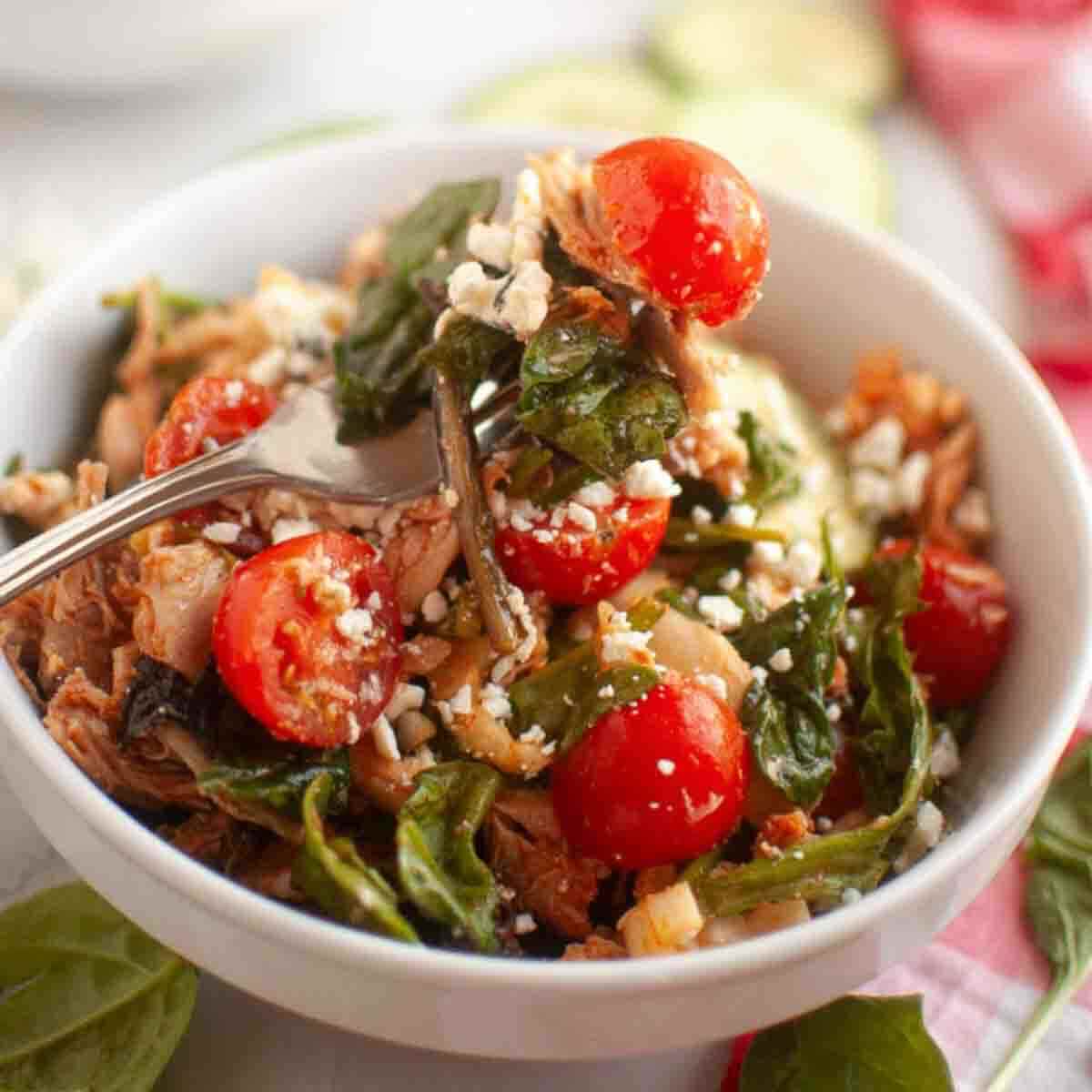 A bowl of salad with shredded meat, cherry tomatoes, spinach, and crumbled cheese, with a fork resting inside.