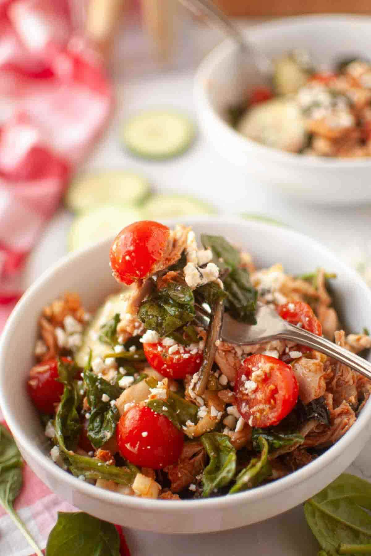 A bowl of rice mixed with cherry tomatoes, spinach, shredded meat, and crumbled cheese, with a fork resting inside.
