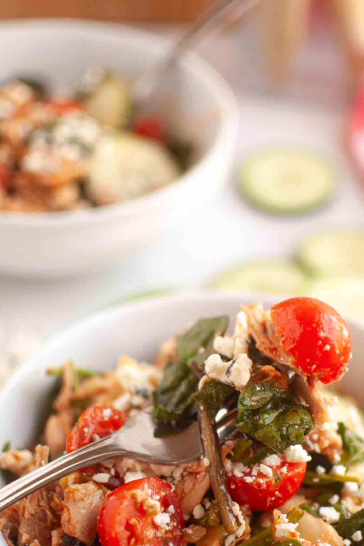 A close-up of a fork holding a bite of salad with cherry tomatoes, leafy greens, grains, and crumbled cheese, with a bowl of salad in the background.