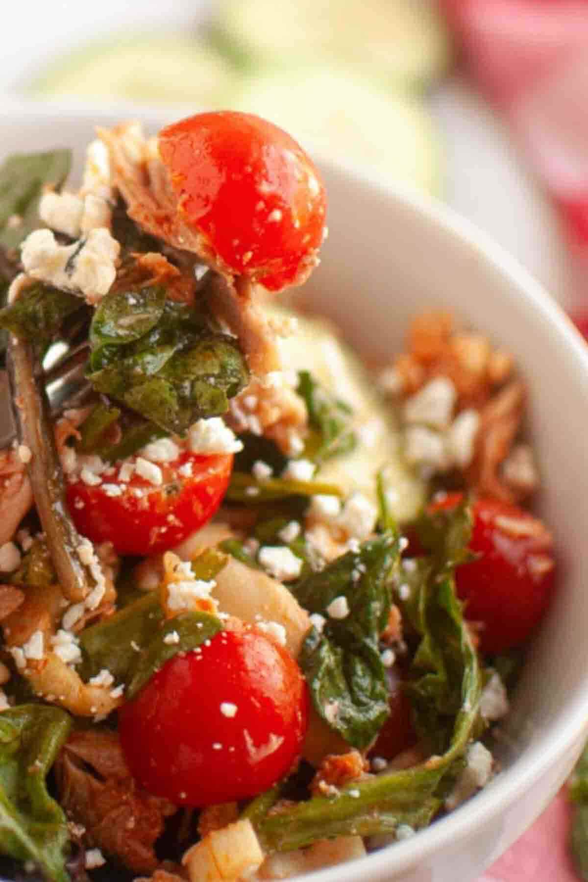 A close-up of a salad with cherry tomatoes, leafy greens, shredded meat, and crumbled cheese in a white bowl.