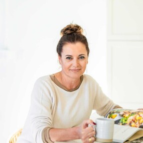 Woman with brown hair in a bun sits at a table holding a mug and looking at the camera, with an open cookbook in front of her.
