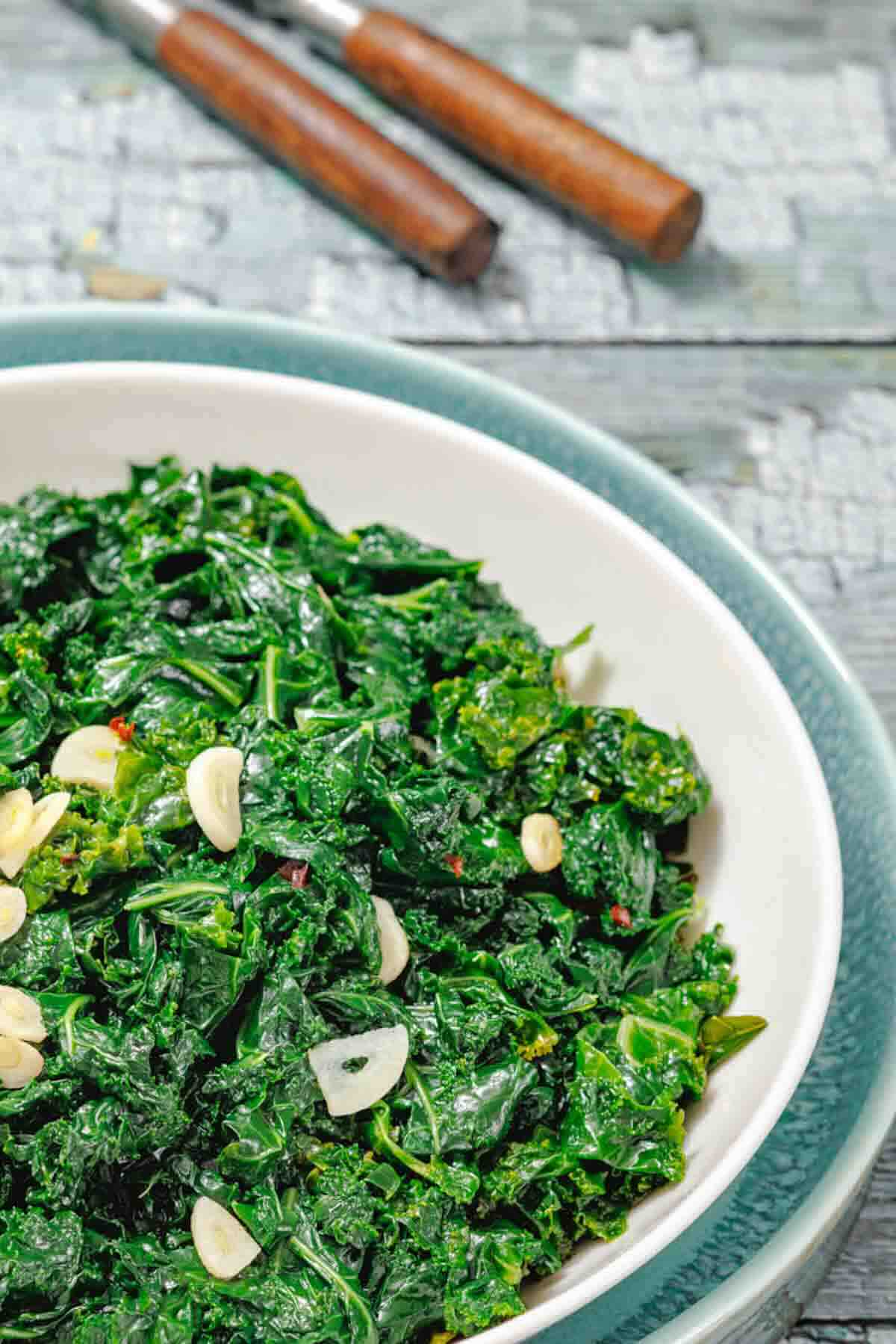 A bowl of sautéed kale with sliced garlic on top, placed on a blue and white plate with two utensils in the background.