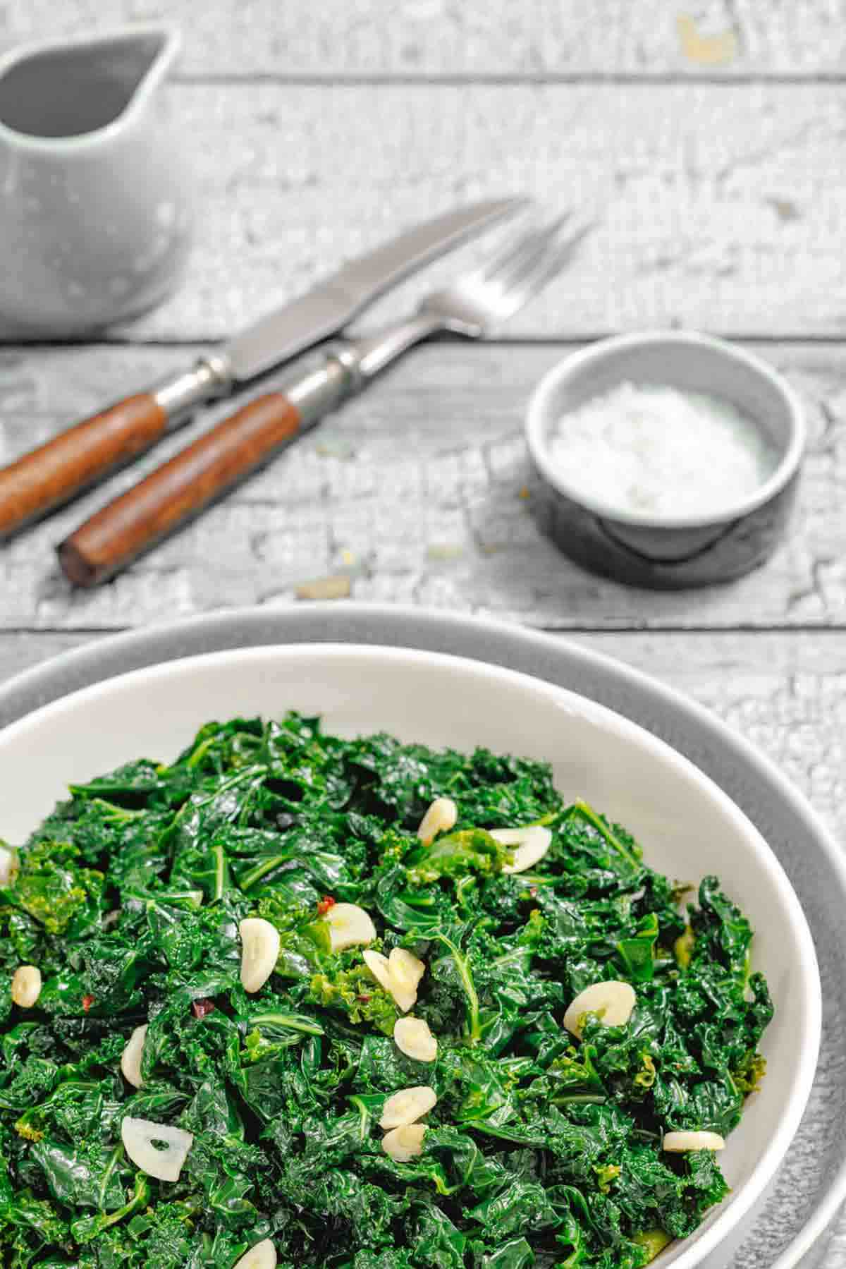 A white bowl filled with sautéed kale and sliced garlic sits on a rustic wooden table, with a small bowl of salt, a fork, knife, and a jug in the background.