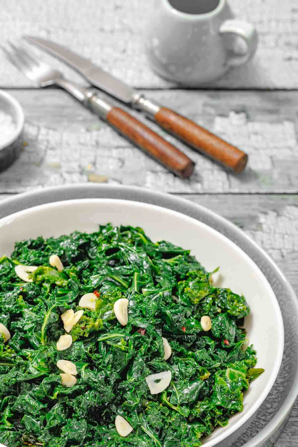 A bowl of sautéed kale topped with sliced garlic, placed on a plate with a fork and knife in the background.
