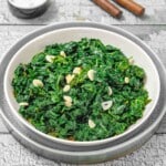 A white bowl filled with sautéed kale and sliced garlic, placed on a plate on a rustic wooden surface.
