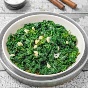 A white bowl filled with sautéed kale and sliced garlic, placed on a plate on a rustic wooden surface.
